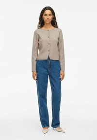 Cardigan beige clair à manches longues avec des boutons en or, associé à un jean bleu décontracté. Le tissu semble doux avec une texture lisse.