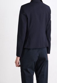 Person sedd bakifrån iförd en skräddarsydd mörk marinblå kavaj och matchande mörk marinblå byxor mot en enkel ljus bakgrund.