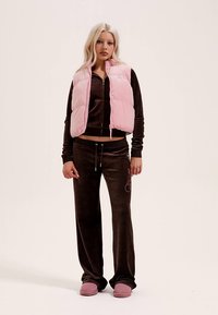 Różowa pikowana kamizelka, brązowa aksamitna kurtka na zamek, dopasowane spodnie z różowym logo oraz różowe kapcie. Miękkie tekstury, luźny krój.