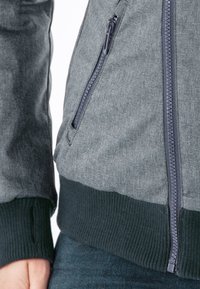 Graue Textil-Bomberjacke mit einer Reißverschlusstasche, gerippten Bündchen und einem kontrastierenden dunklen gerippten Saum. Glatte Stoffoberfläche mit einem dezenten Muster.