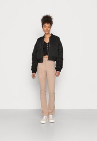 Jaqueta bomber cropped preta com punhos canelados, combinada com calças de cintura alta beje risca e ténis brancos, criando uma silhueta ajustada.