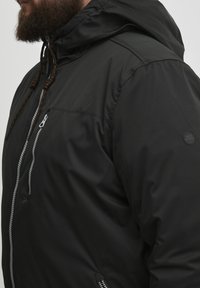 Sort isoleret jakke med hætte, der har en frontlynlås, en brystlomme og orangerøde snore. Glat, vandafvisende stof.