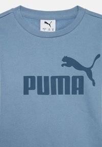 T-shirt en coton bleu clair avec un grand logo "PUMA" bleu marine et une silhouette de puma sautant au-dessus du texte. Design à col rond.