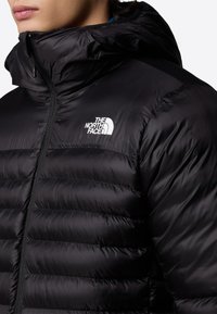 Zwarte pufferjack met hoge kraag, ritsluiting en het logo van The North Face op de linkerborst. Heeft een gestikte, gevoerde structuur.