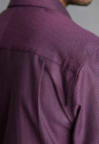 Nahaufnahme einer Person, die ein lilafarbenes Hemd mit einem feinen schwarzen Fischgrätmuster auf der Schulter und dem Ärmel trägt.