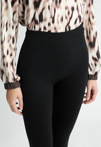 Leggings negros con una alta cinturilla, hechos de un material elástico. La cinturilla presenta una textura acanalada. Blusa visible con un patrón de manchas.