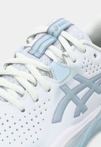 Hvit atletisk sko med perforert overdel, lyseblå detaljer og teksturert logo. Den har flate lisser og en polstret såle.