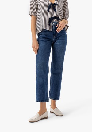 Osoba nosząca niebieskie dżinsy z wysokim stanem i prostymi nogawkami, pasiasty top z krótkim rękawem ozdobiony granatowymi kokardami oraz białe mokasyny na białym tle.