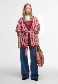 Κόκκινη και ροζ καρό κάρντιγκαν με ζώνη, συνδυασμένη με μπλούζα με τύπωμα και φαρδιά τζιν παντελόνια. Κρατάει μια τσάντα με σχέδιο και πράσινες λεπτομέρειες.