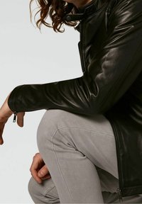 Giacca di pelle nera con un polsino con zip, texture liscia e design aderente, abbinata a pantaloni grigio chiaro con dettagli a righe.