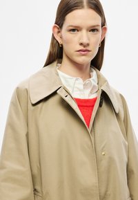 Bézs, könnyű ballonkabát klasszikus gallérral, elülső cipzárral és arany részletekkel, élénk piros pulóver és fehér ing fölött viselve.