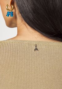Frau mit langen, dunklen Haaren, die goldene Creolen mit blauen Perlen und einen gerippten goldenen Pullover mit einem kleinen metallischen Bienen-Detail auf dem Rücken trägt.