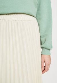 Felpa verde chiaro abbinata a una gonna a pieghe color crema. La gonna presenta pieghe verticali e una fascia elastica in vita.