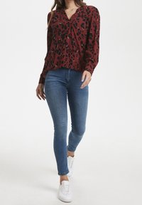 Rote Leopardenmuster-Bluse mit V-Ausschnitt, langen Ärmeln und lockerer Passform, kombiniert mit eng anliegenden blauen Jeans und weißen Turnschuhen.