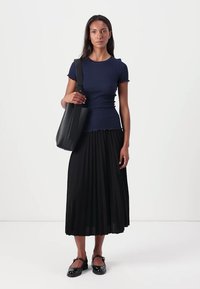 Laivaston sininen muotoiltu toppi, jossa on aaltoileva helma, pleated musta midi-hame ja mustat slingback-kengät. Malli pitää suurta mustaa laukkua.