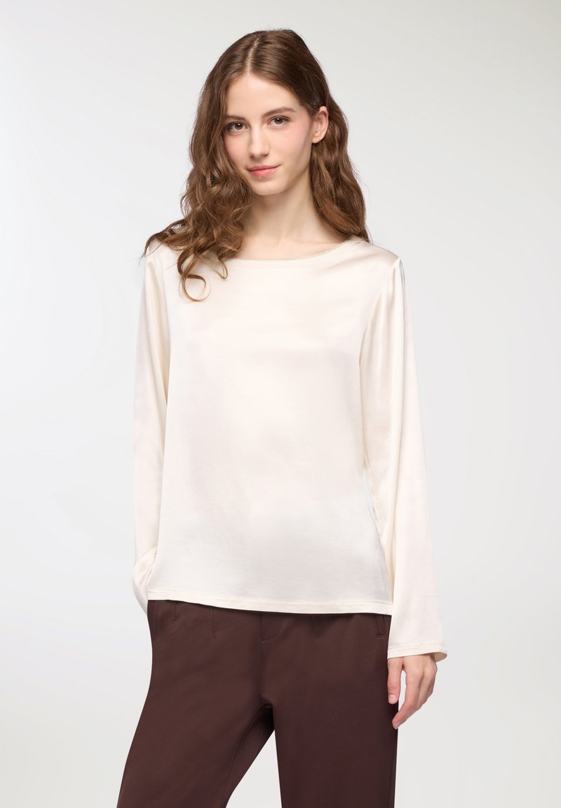 Blusa a maniche lunghe color crema con scollatura a barchetta, morbida, di texture liscia, fitted leggermente largo, abbinata a pantaloni marrone scuro.