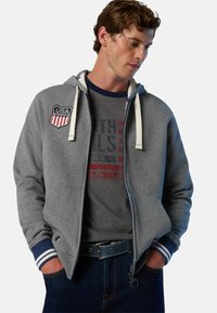 Felpa grigia con zip e lacci bianchi, con una patch degli Stati Uniti. Sotto indossa una maglietta nera con testo grafico rosso e blu. Sono visibili dei jeans blu.