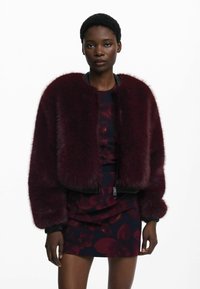 Chaqueta bomber de pelo sintético burdeos con puños y cuello acanalados en negro, con cierre de cremallera en la parte delantera y diseño corto, que complementa un vestido oscuro con estampado floral.