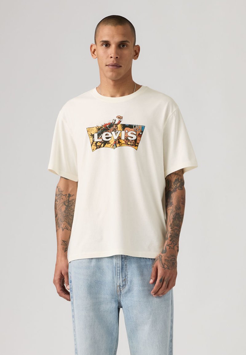 Camiseta de algodón color crema con mangas cortas y un logotipo gráfico en el frente. Se lleva con vaqueros de mezclilla azul claro.