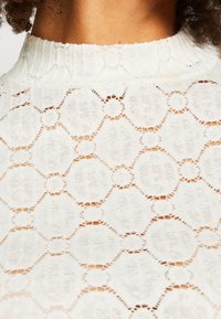 White lace top featuring a textured pattern of interlinked circles and hexagons, with a high neckline and ribbed detailing.