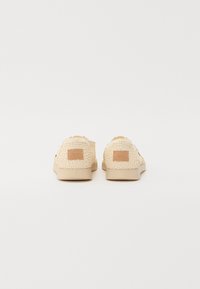 Beige nahkatossut, joissa on kudottu tekstiilipinta, kerma väri pohja ja pieni ruskea logopläntti kantapäässä. Avoin takaosa helpottamassa käyttöä.