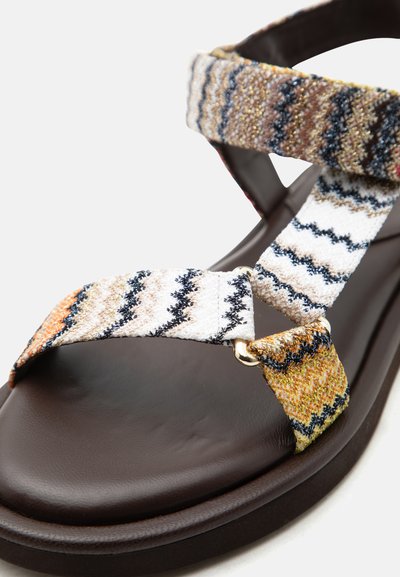 Σανδάλι με ανοιχτά δάχτυλα, σκούρα καφέ σόλα και πολύχρωμες υφασμάτινες λουρίδες με ζιγκ-ζαγκ σχέδιο, στερεωμένες με μεταλλικούς κρίκους σε χρυσό τόνο.