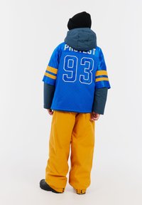 Blaues Netztrikot mit gelben Streifen und dem Text "PROTEST 93", kombiniert mit orangen Hosen und einer schwarzen Mütze, von hinten getragen.