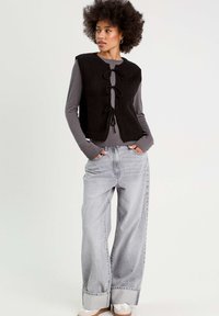 Gilet en tricot noir avec des liens à l'avant, superposé sur une chemise à manches longues grise. Le modèle porte un jean large gris clair et des chaussures crème.