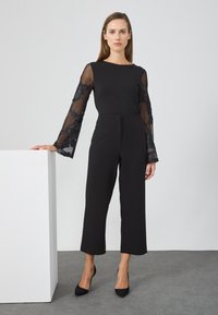 Jumpsuit nero con lunghe maniche in pizzo nero trasparente caratterizzate da modelli floreali. Il design include un taglio dritto e un collo alto.