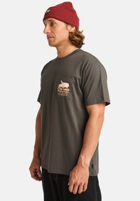 Camiseta gris de manga corta con un bolsillo en el pecho que presenta una ilustración y un diseño de texto. El modelo lleva un gorro rojo.