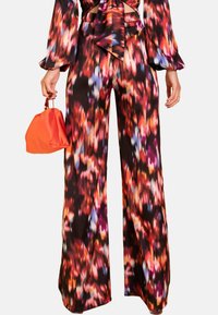 Pantalones anchos y largos con un estampado floral en vibrantes rojos, púrpuras y azules sobre un fondo negro; combinados con un pequeño bolso naranja.