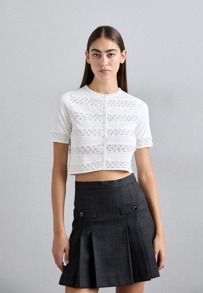 Young woman stands against a plain gray wall wearing a white short-sleeve pearl-embellished cropped top and dark pleated plaid skirt.