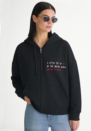 Vrouw met steil zwart haar, een zonnebril, een zwarte zip-up hoodie met tekst en lichtblauwe spijkerbroek tegen een neutrale achtergrond.