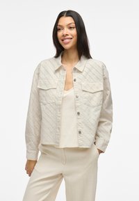Lys beige kort jakke med diagonalt teksturmønster, to brystlommer, trykknappestengning og lange ermer, stylet over en nøytral topp.
