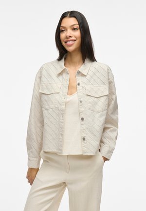 Light beige cropped jacket with diagonal textured pattern, two chest pockets, snap closures, and long sleeves, styled over a neutral top.