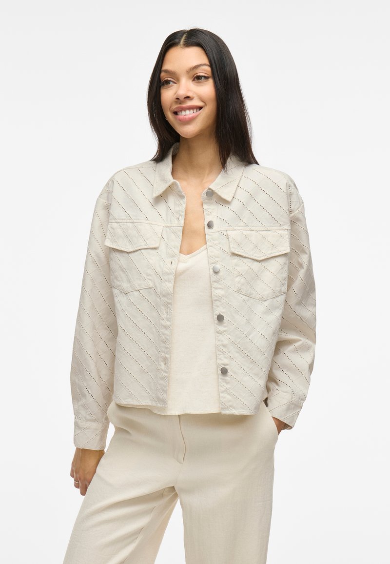 Lys beige kort jakke med diagonalt teksturmønster, to brystlommer, trykknappestengning og lange ermer, stylet over en nøytral topp.