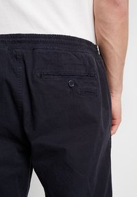 Lähellä otettu kuva henkilöstä, joka on pukeutunut mustiin elastisiin vyötäröhousuihin, joissa on kaksi takataskua, joista toinen on napitettu, ja näkyy osa valkoisesta paidasta ja käsivarresta.