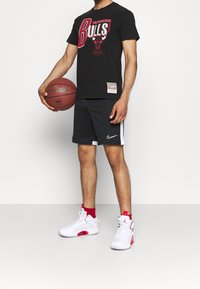 Zwart graphic t-shirt van de Chicago Bulls, zwarte shorts met witte zijstrepen, en wit/rode basketball schoenen. Model houdt een basketbal vast.