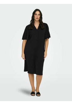 Kvinde med langt mørkt hår iført en løs sort knælang kjole med korte, brede ærmer og sorte åbne sko, stående mod en ensfarvet baggrund.