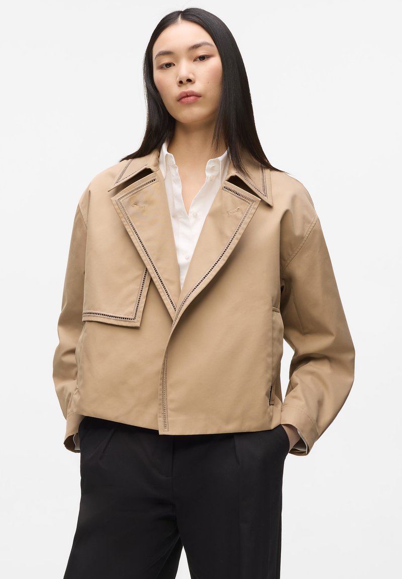 Νεαρή γυναίκα με μακριά μαύρα μαλλιά φοράει μπεζ κοντό σακάκι πάνω από λευκό πουκάμισο με κουμπιά και μαύρο παντελόνι, με τα χέρια στις τσέπες, μπροστά σε απλό φόντο.