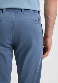 Helle blaue Hose aus glattem Stoff; Merkmale sind Gürtelschlaufen, zwei Gesäßtaschen und ein dezentes Markenschild nahe der rechten Tasche.