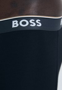Schwarze Boxerbriefs mit einem breiten elastischen Bund, der das weiße „BOSS“-Logo und einen kontrastierenden grauen und goldenen Streifen aufweist. Glatte Textur.