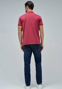 Hombre de pie de espaldas a la cámara, vistiendo una camiseta polo roja, jeans azules y zapatillas blancas, frente a un fondo gris liso.