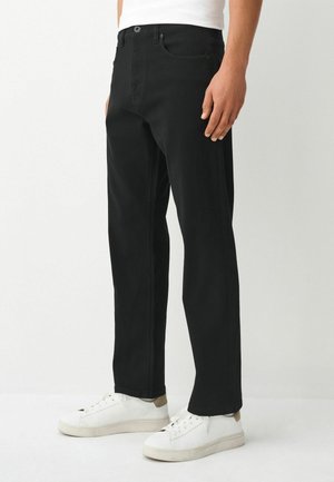 Mann trägt schwarze gerade geschnittene Hose und weiße Sneakers, steht vor einem einfarbigen hellen Hintergrund.