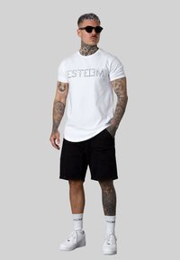 Weißes Baumwoll-T-Shirt mit schwarzem Drucktext, kombiniert mit schwarzen Shorts. Getragen mit weißen Turnschuhen und dunklen Sonnenbrillen, mit auffälligen Tattoos.