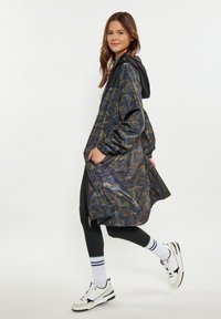 Waterdichte jas met camouflagedesign in marineblauw en groene tinten. Heeft een capuchon, elastische manchetten en een relaxte pasvorm, gedragen over zwarte leggings.