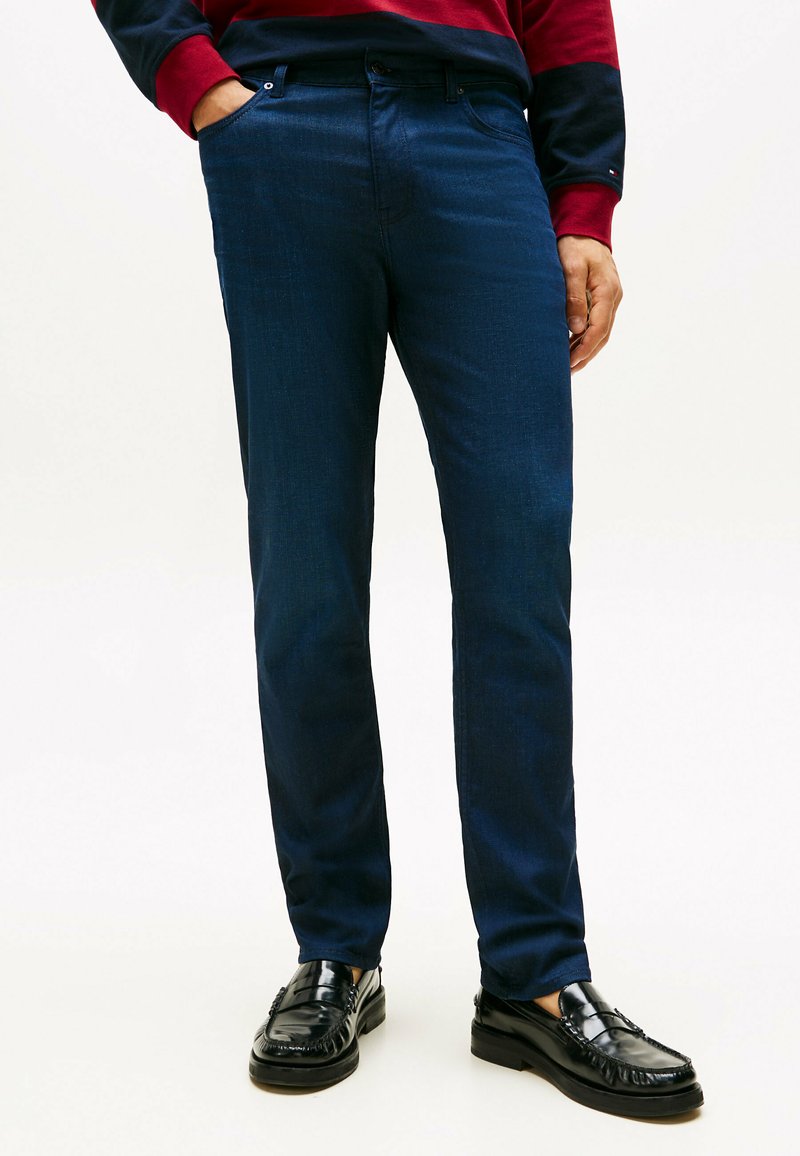 Hombre vistiendo jeans azul oscuro, mocasines de cuero negros y una camisa de manga larga roja y azul marino, con una mano en el bolsillo.