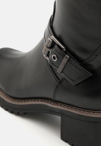 Stivale alla caviglia in pelle nera con suola piatta, dotato di una fibbia decorativa in metallo, texture liscia e dettagli di cuciture a contrasto.