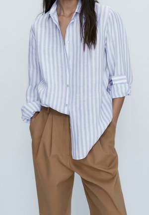 Femme portant une chemise blanche à rayures verticales bleu clair, manches retroussées, rentrée dans un pantalon plissé marron taille haute.