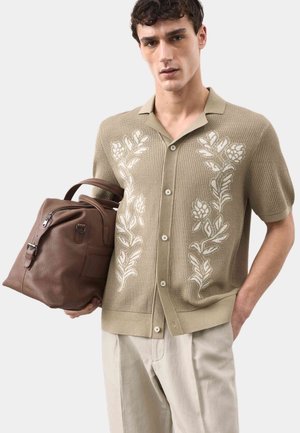 Junger Mann trägt beige kurzärmliges Strickshirt mit Blumenstickerei, hält eine große braune Ledertasche, steht vor neutralem Hintergrund.
